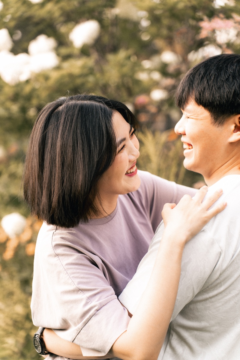 A man and woman share a candid kiss and hug outdoors, with the woman looking happy and trees in the background.