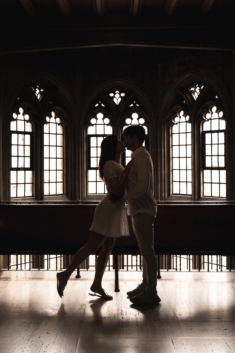 A silhouetted couple dances indoors in front of a window as the girl leans in for a kiss, captured in black and white.