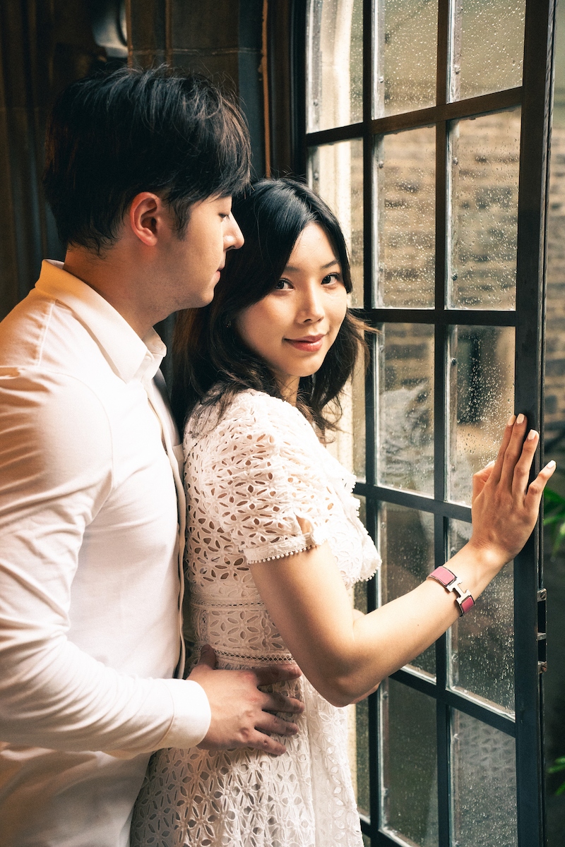 A man embraces his partner, who smiles at the camera while he looks intimately at her. She's in a wedding dress, and they stand indoors by a window.