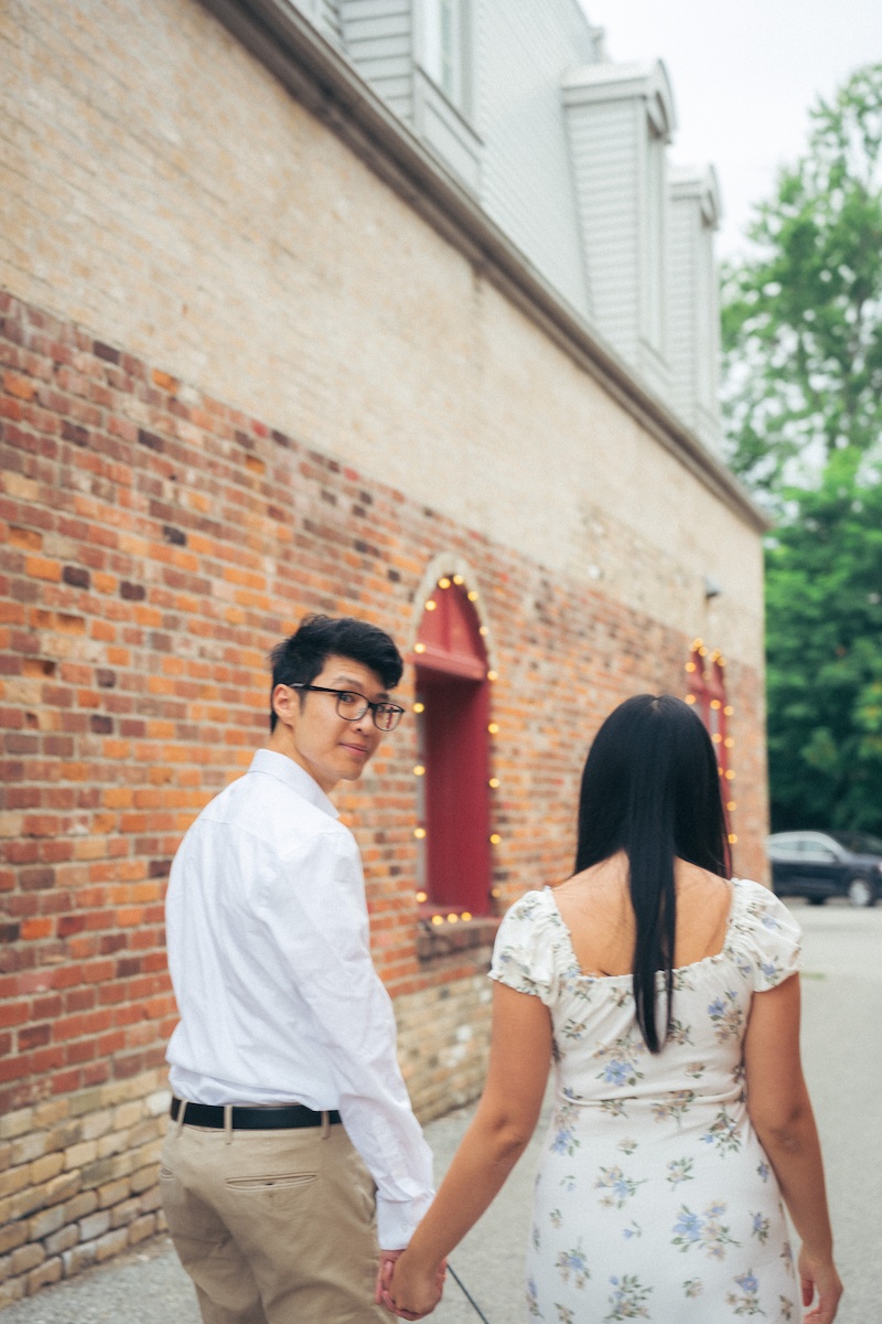 A couple holds hands while walking down a red-brick alley. The man wears a casual summer outfit with glasses, and the woman is in a summer dress.