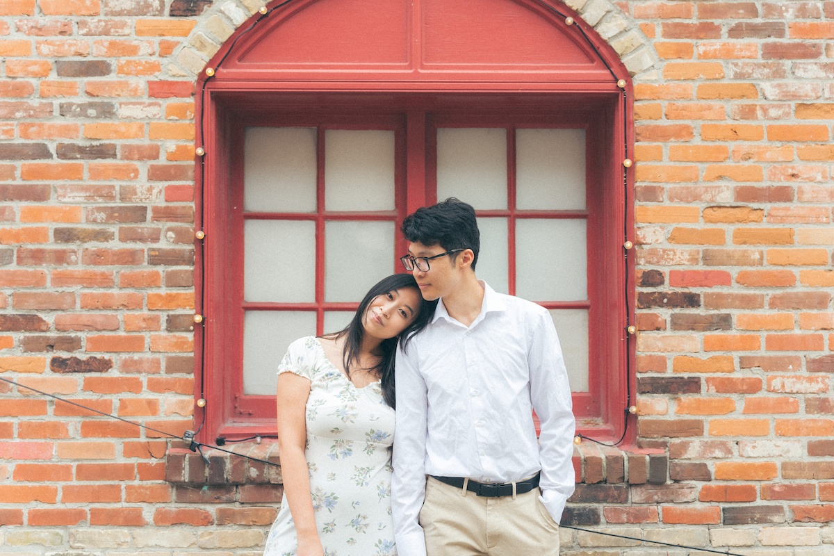 A man and woman pose in front of a red window, dressed for an engagement occasion, with brick walls and a window in the background. Girl is leaning on partner's shoulder