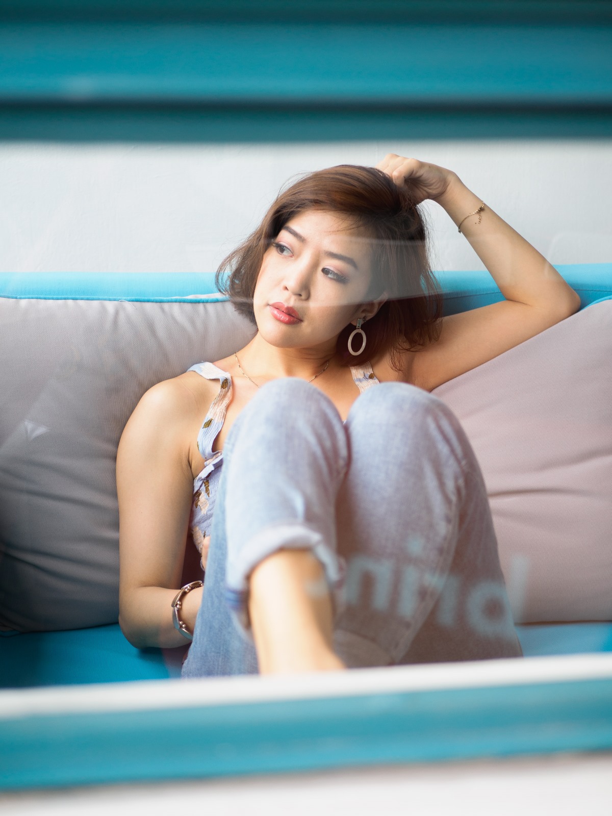 An asian woman in deep thought while lying on a couch. She has short hair and is wearing blue jeans.
