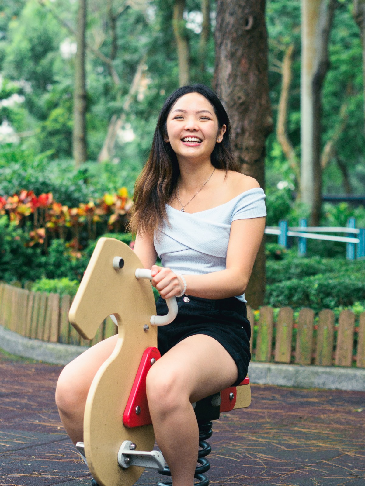 A smiling asian woman discovers her childhood and playfully rides a tricycle in a park, wearing fashionable clothing, with trees and flowers in the background.