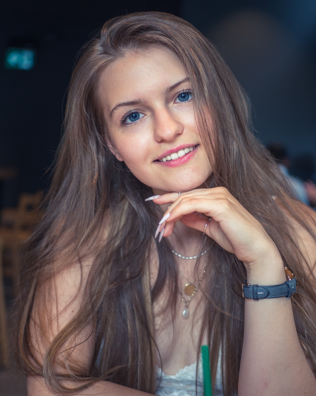 A smiling caucasian woman with long, layered brown hair and lip gloss, posing for a fashion photo shoot in a coffee shop.