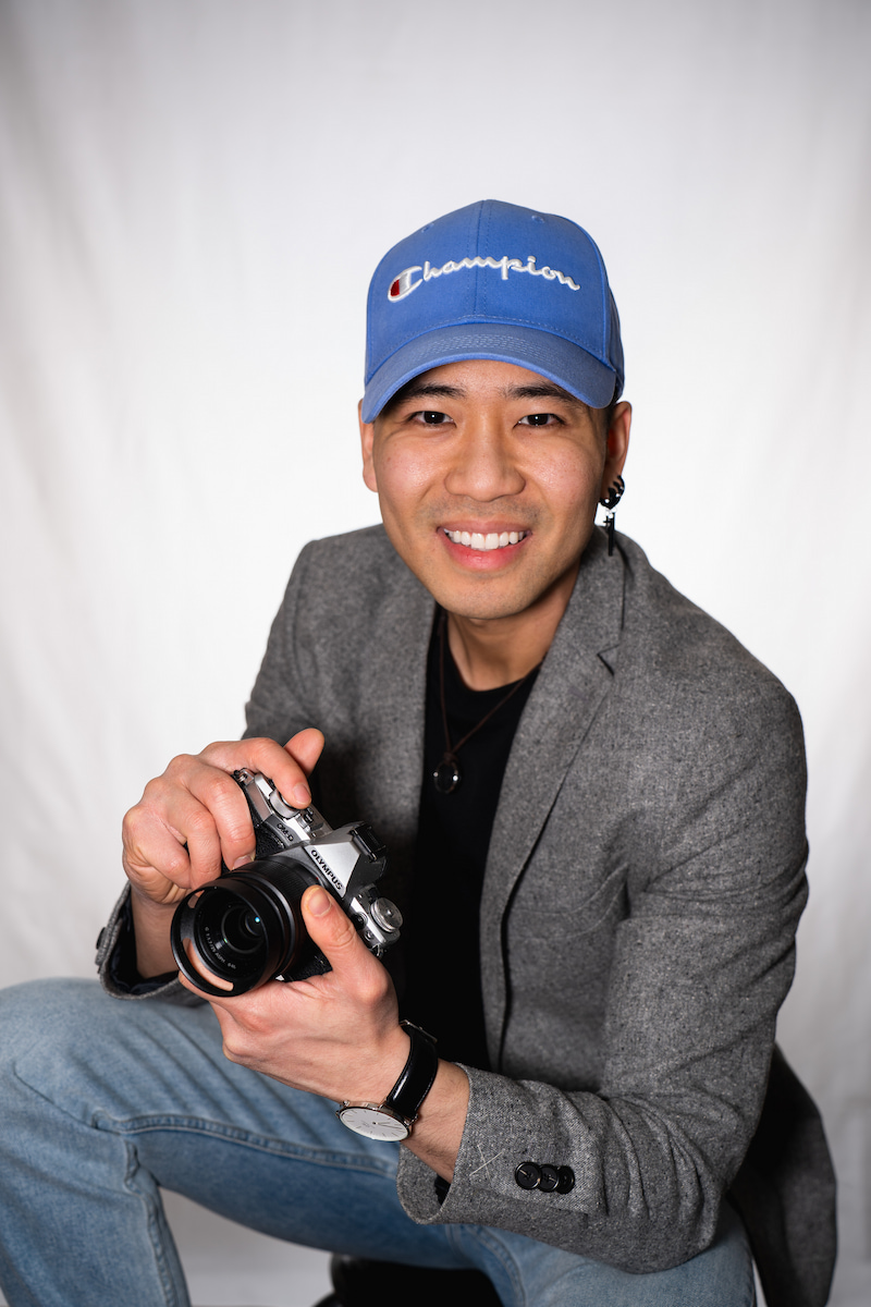 A smiling young Asian man holds a camera while standing on a street, with a vehicle in the background.